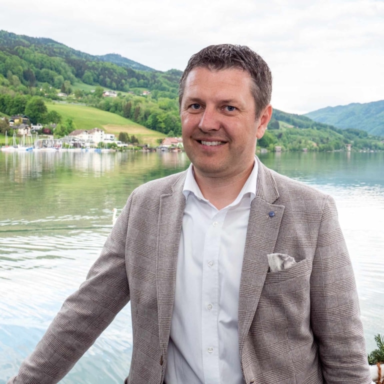Geschäftsführer der Schifffahrt Meindl vor dem Mondsee: Mann im karierten Blazer mit See, Hügeln und Ortschaft.