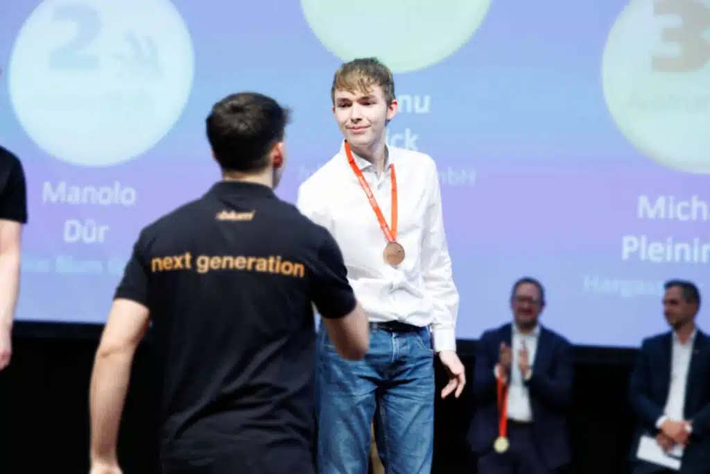 Hargassner-Mitarbeiter Michael bei der Bronzemedaillen-Überreichung auf der Austrian-Skills-Bühne 2025; im Hintergrund die Podestanzeige mit Hargassner auf Platz drei.